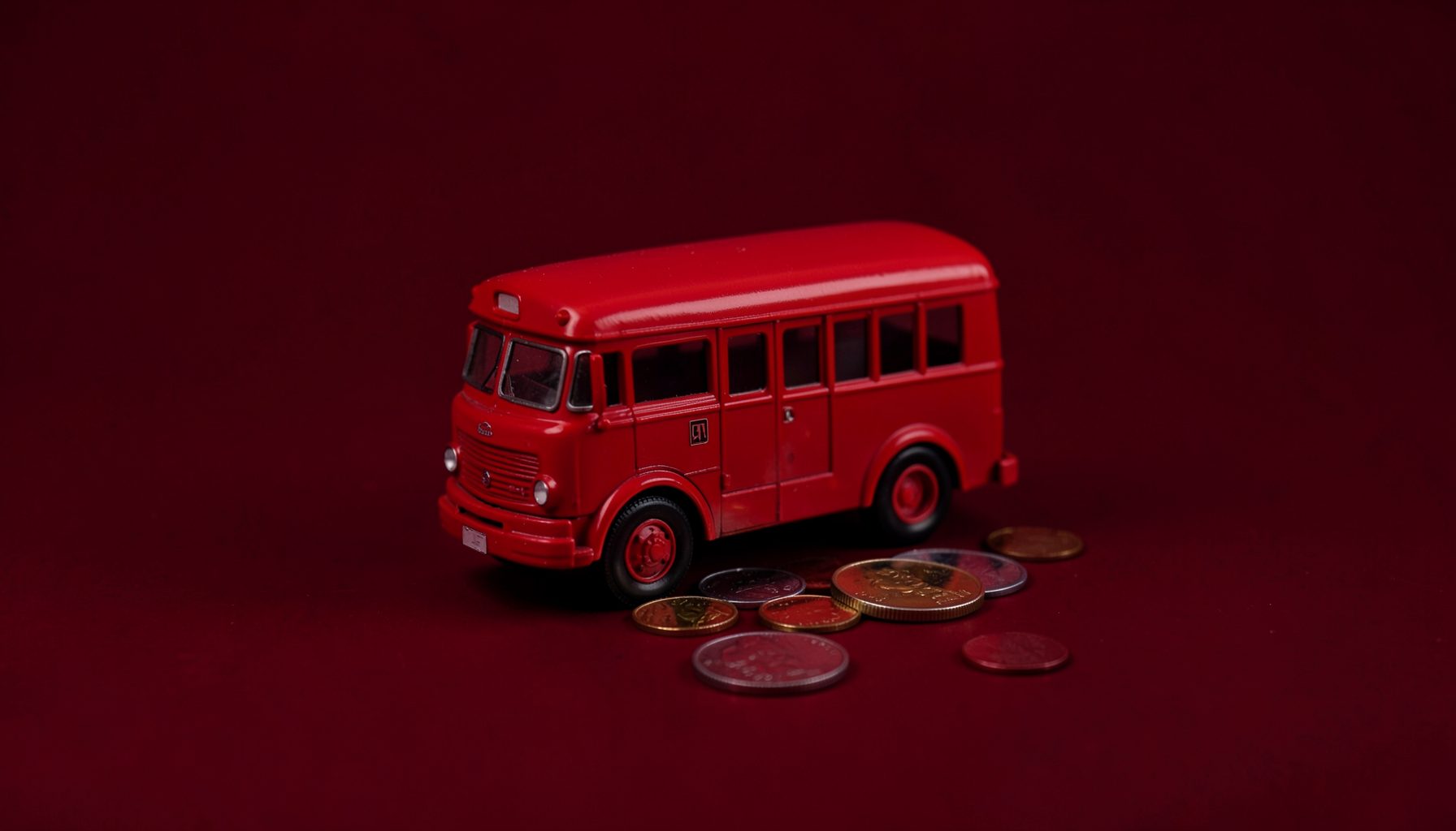 A studio photograph of a red toy bus and coins on a plain black backdrop symbolizing transport fares